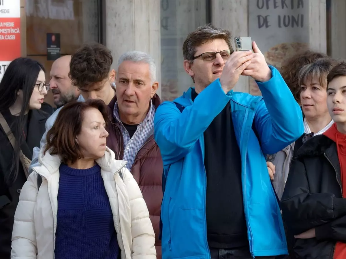 A man in a blue jacket stands in a crowd, looking at his phone. Other people in the crowd are looking at him or at their own phones.