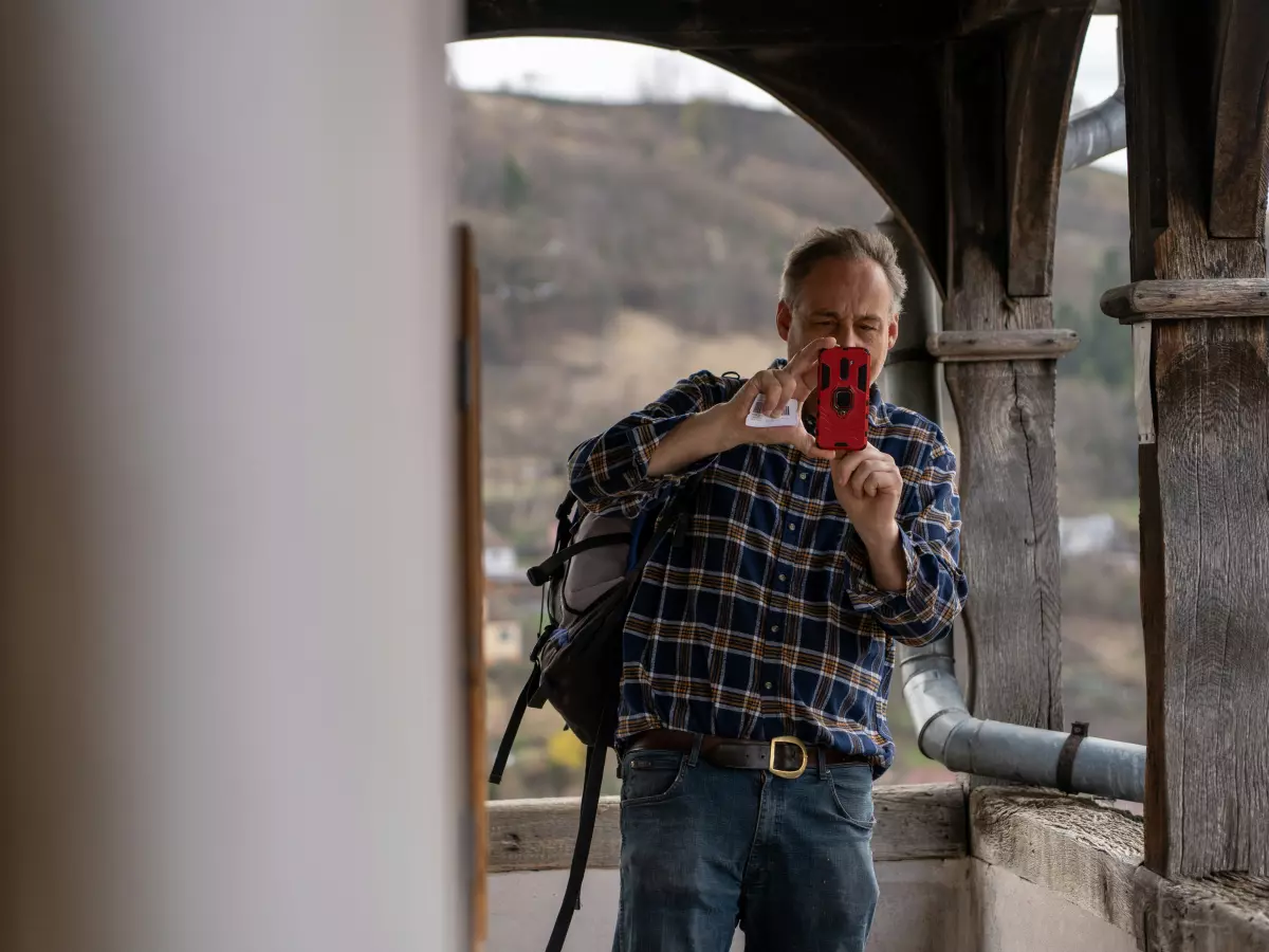 A person is taking a picture with a smartphone. The person is wearing a plaid shirt and has a backpack on their shoulder. The person is standing on a balcony with a view of a forest and a small town in the distance.