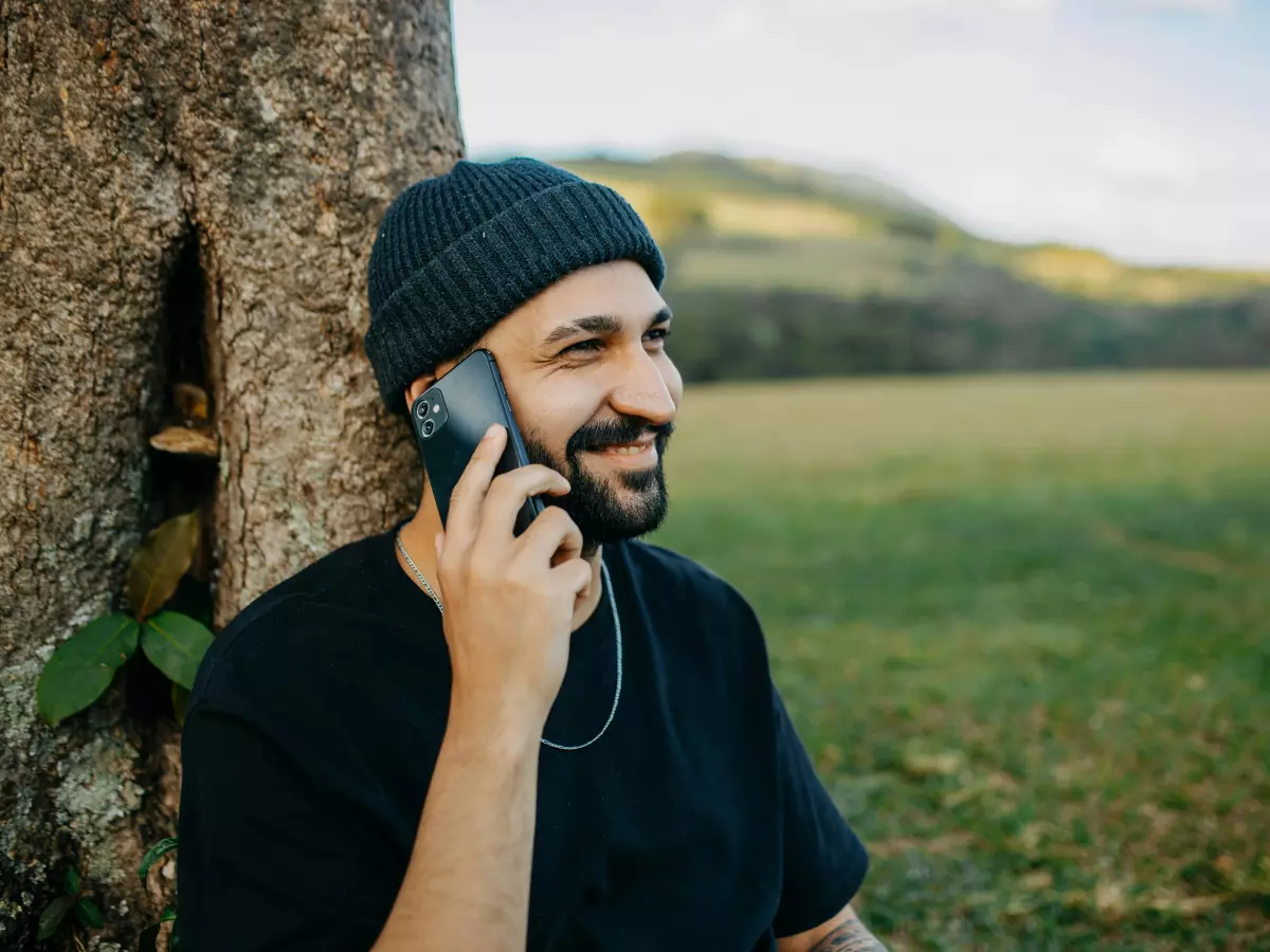 A young man is sitting on a grassy hill with a smartphone in his hands. He is looking at the phone with a slight smile.