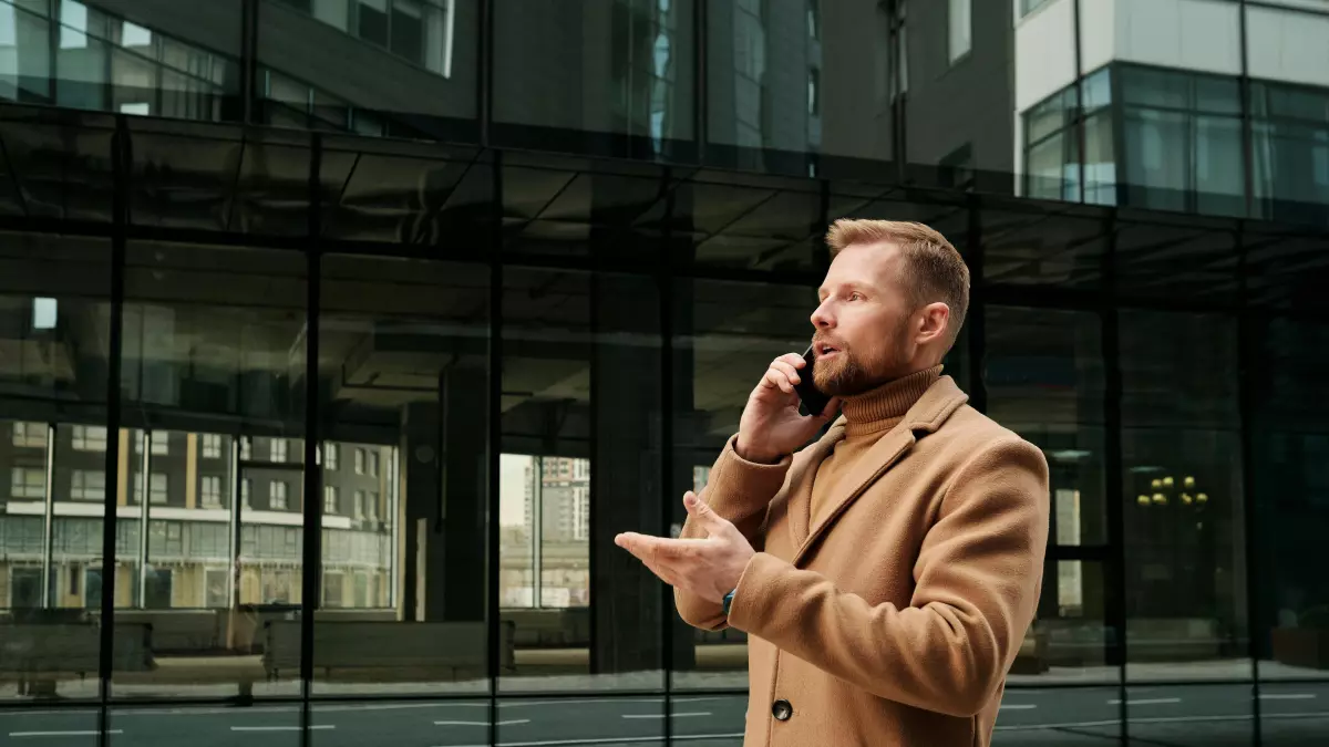 A man in a brown coat talking on a phone in front of a modern building.