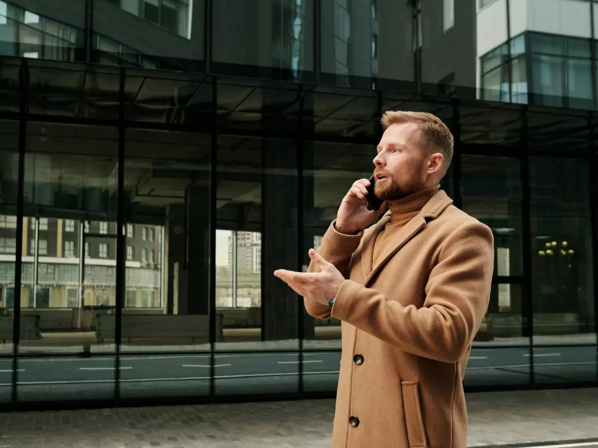 A man in a brown coat talking on a phone in front of a modern building.