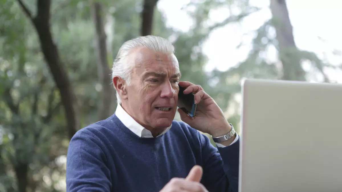 An older man using a laptop and talking on a cellphone.