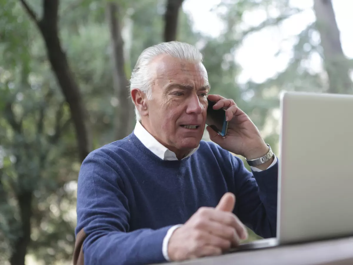 An older man using a laptop and talking on a cellphone.