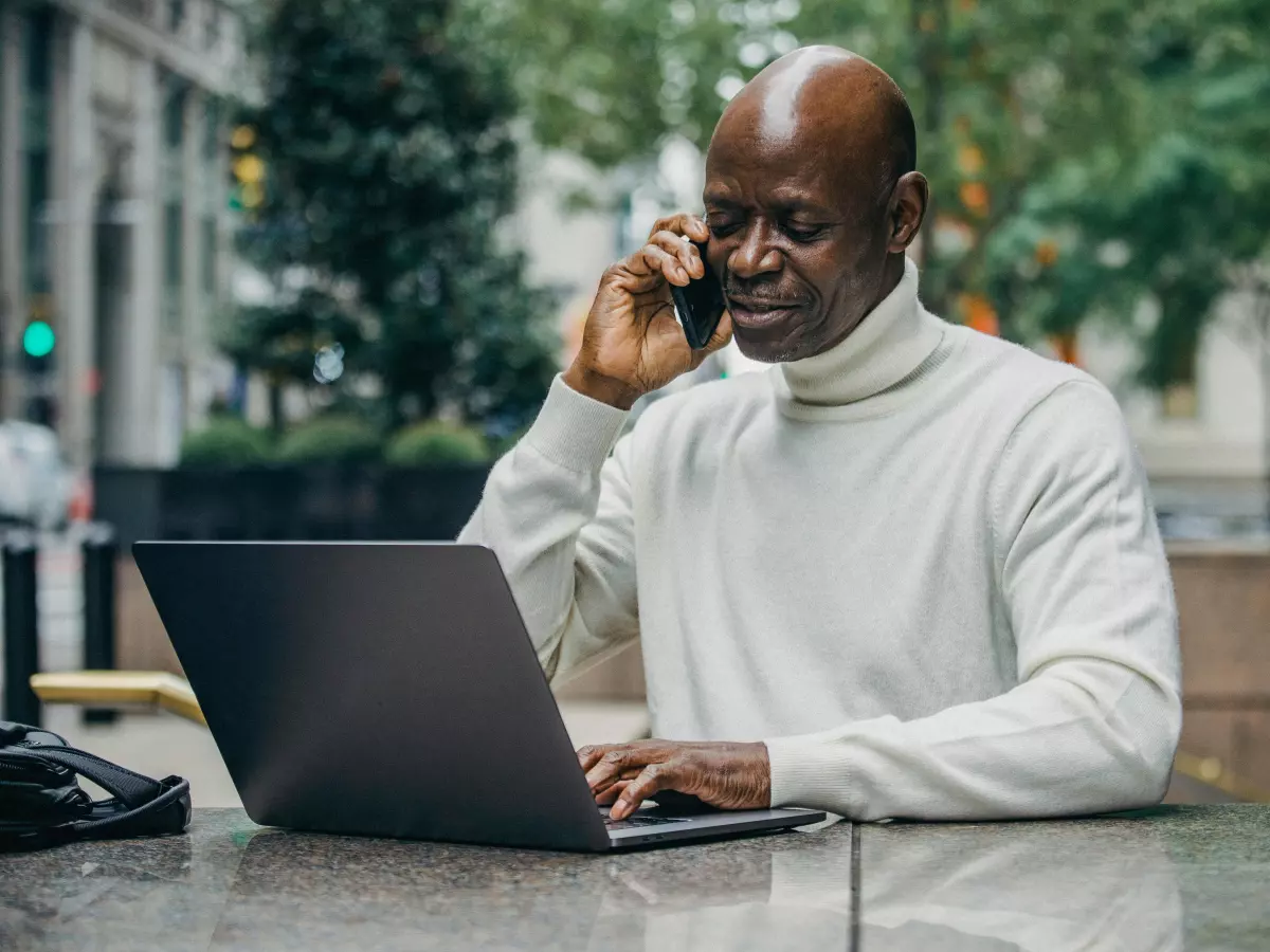 A man wearing a white turtleneck sweater is sitting outdoors, working on his laptop and talking on his phone.