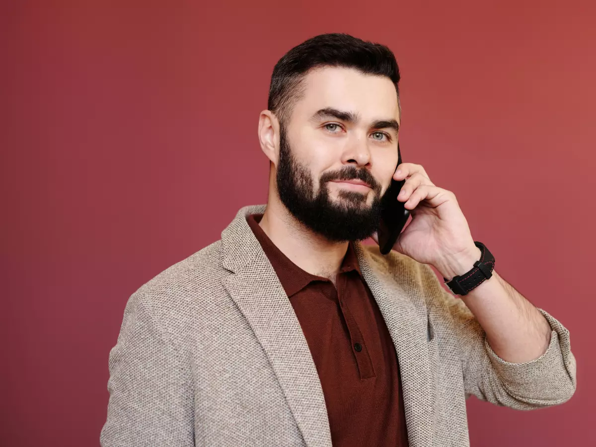 A young man with a beard is talking on his smartphone. He is wearing a beige jacket and a red shirt.