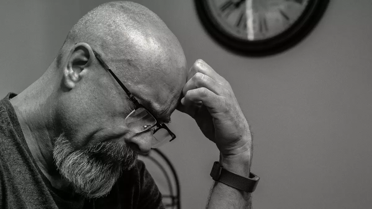 A man with a beard and glasses is looking down, his hand on his chin, in a contemplative pose.