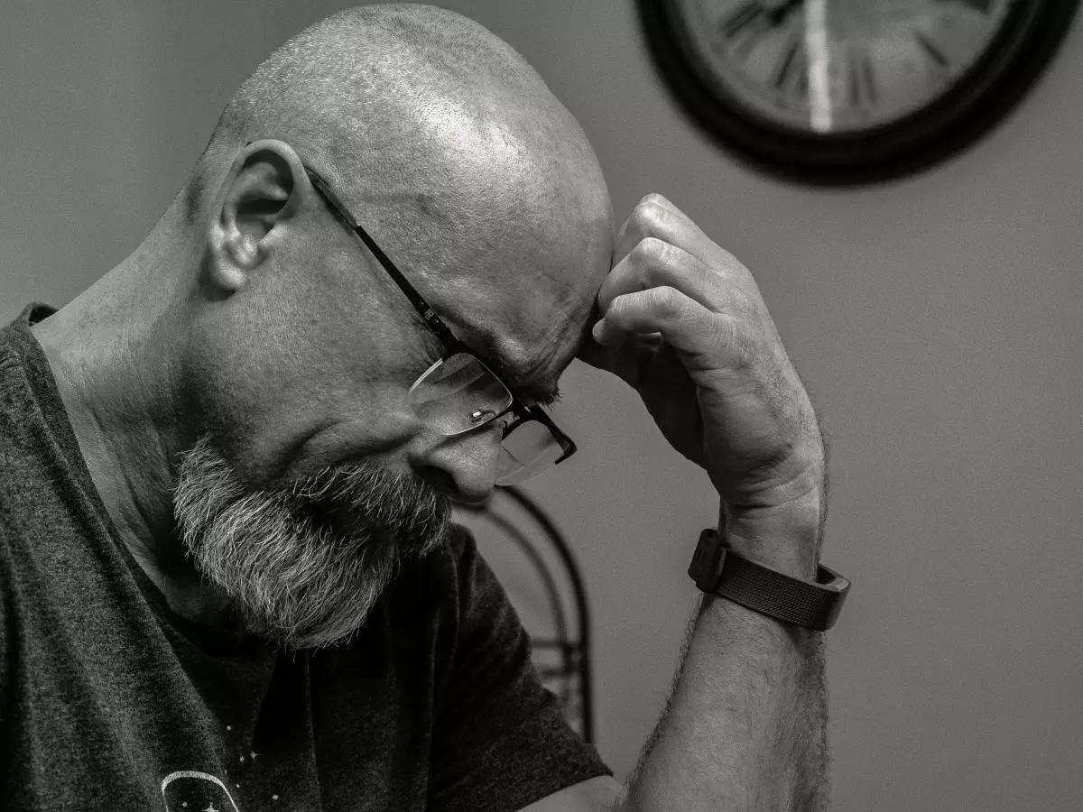 A man with a beard and glasses is looking down, his hand on his chin, in a contemplative pose.