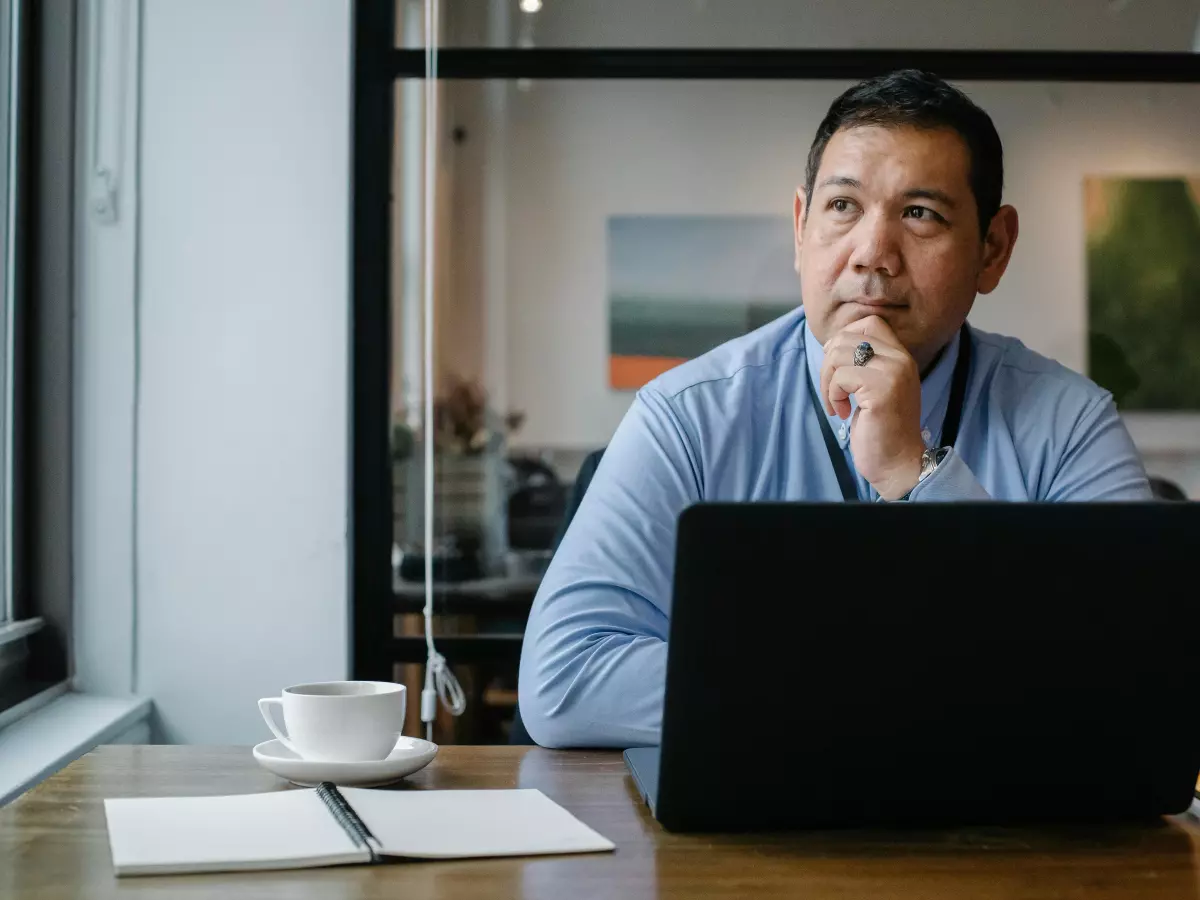 A person is sitting at a table with a laptop in front of them. They are looking off to the side and have a thoughtful expression on their face.