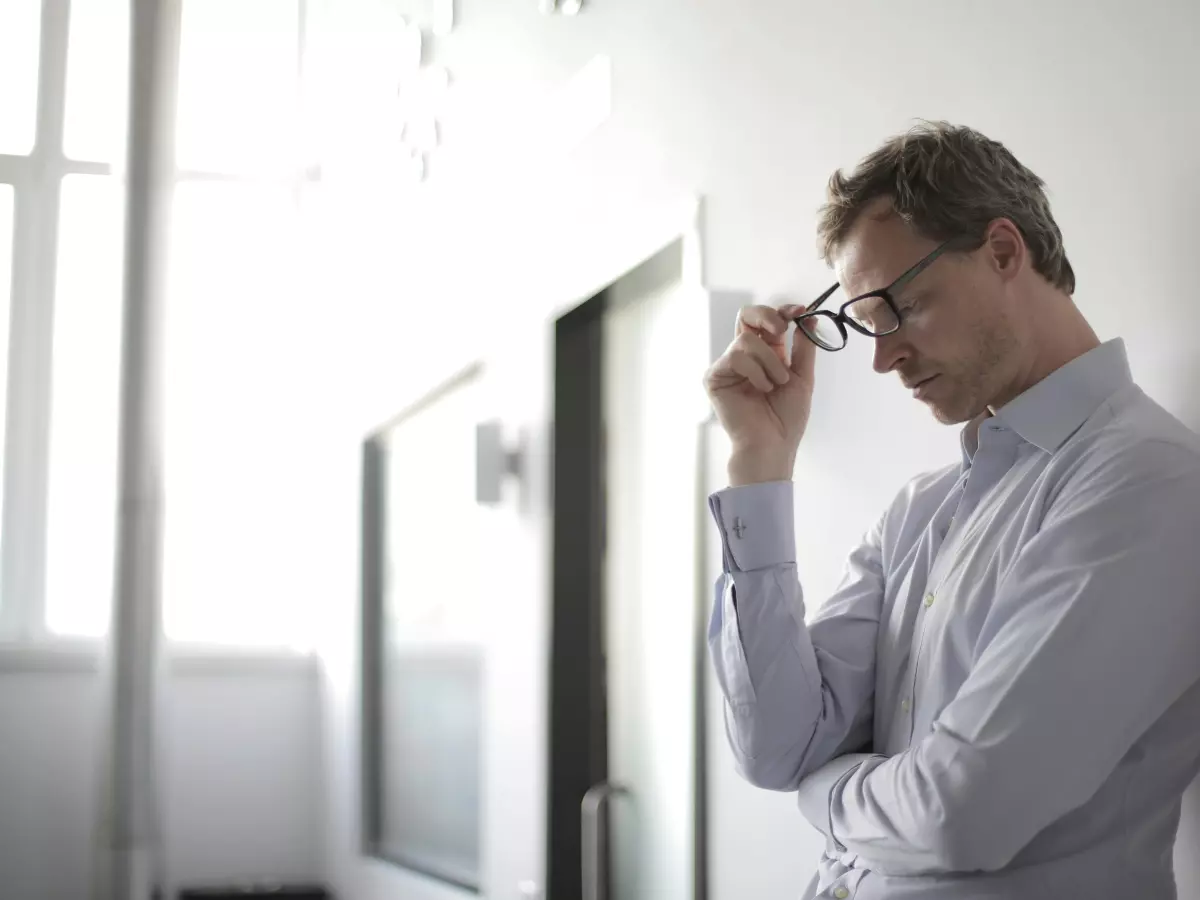 A man in a white shirt and glasses stands against a wall, looking down and holding his glasses with one hand, the other hand on his chin. He is in a state of contemplation.