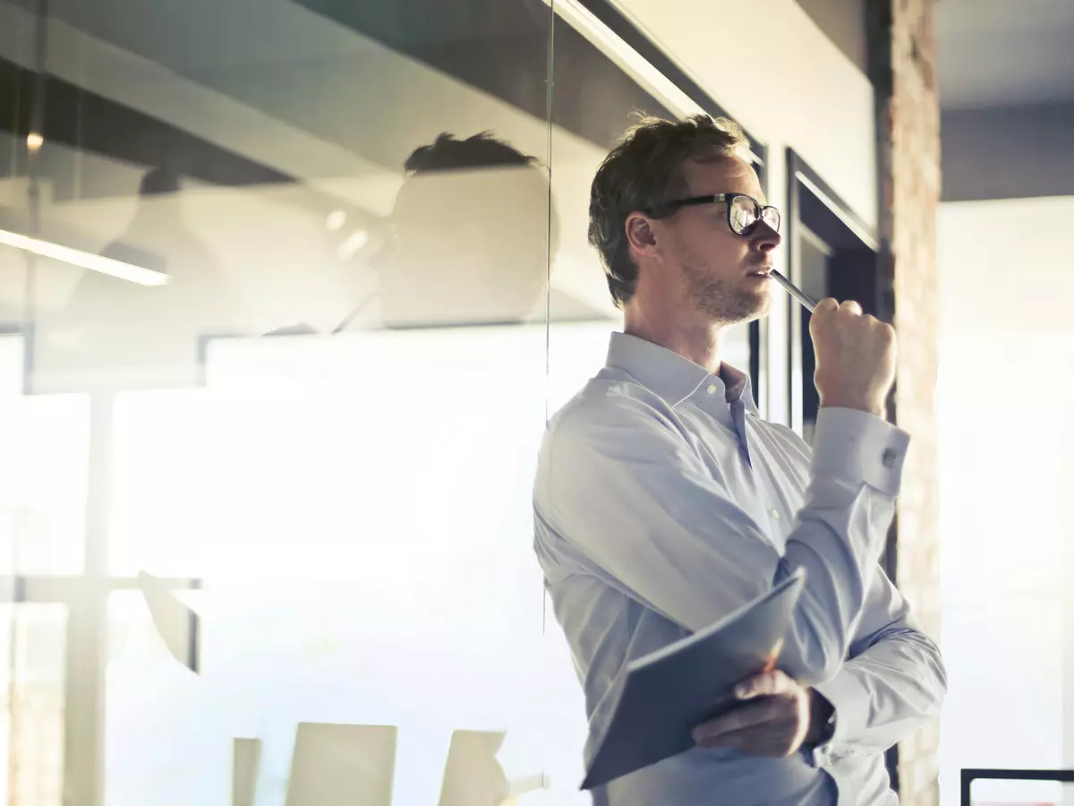 A man in a light blue shirt and glasses stands in an office setting, looking out a window. He has his hand on his chin and is in a pensive pose. The background is blurred, giving the image a sense of depth and solitude.