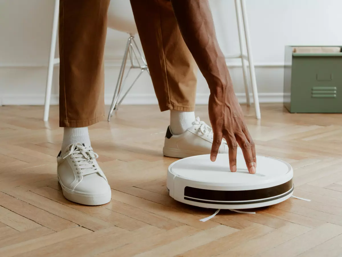 A close-up of a person's hand interacting with a white round robot vacuum cleaner on a wooden floor.