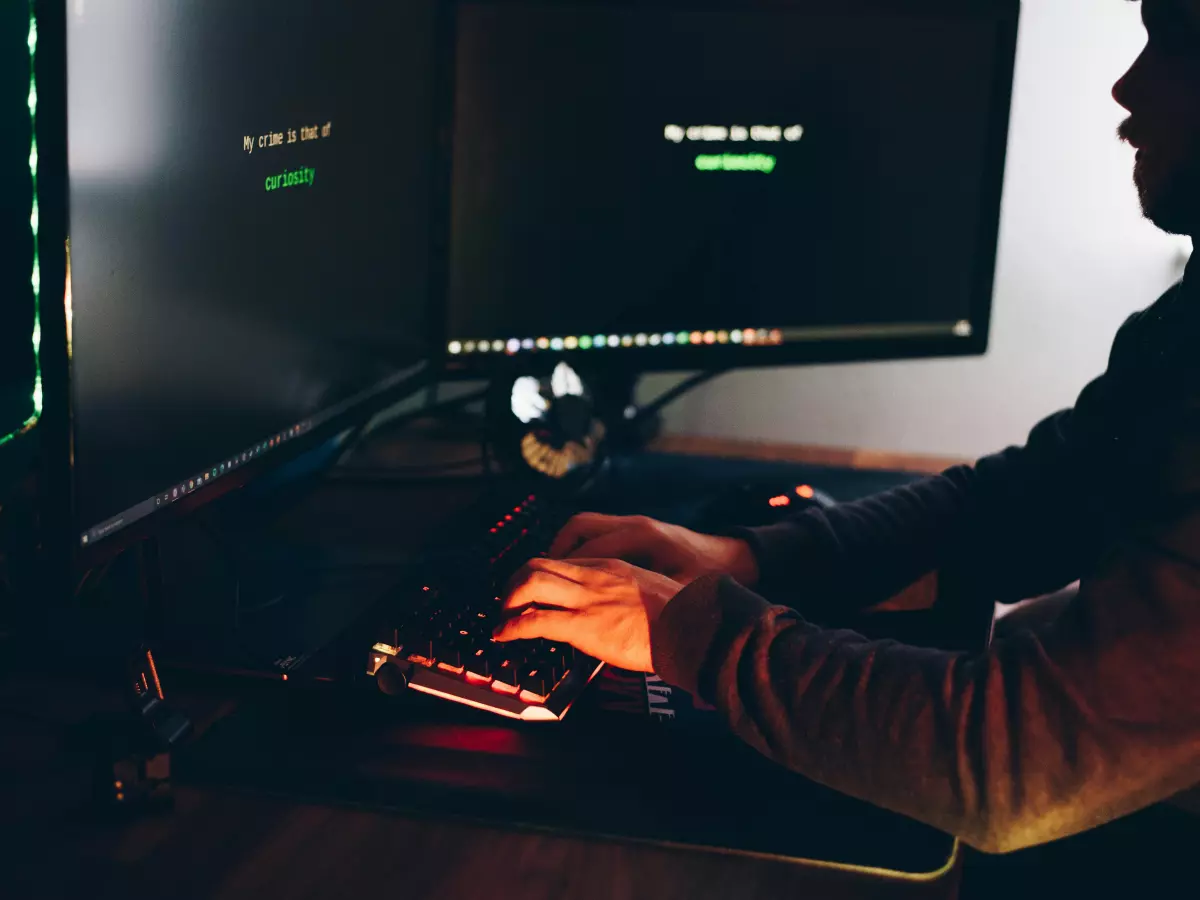 A person working on a computer with green code on the screen. The image is dark and moody, with the person's face obscured.