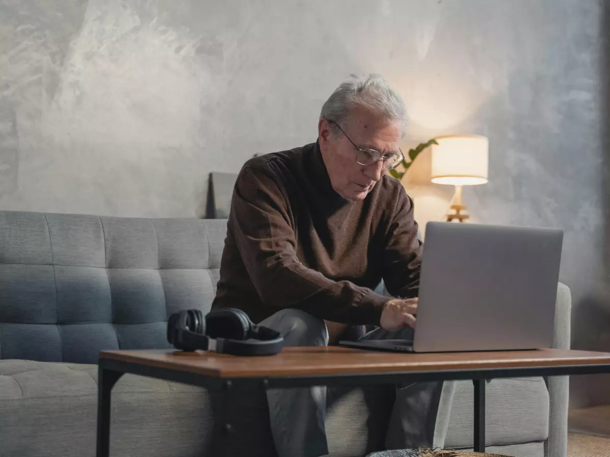 A senior man is sitting on a couch, looking at his laptop with a worried expression.