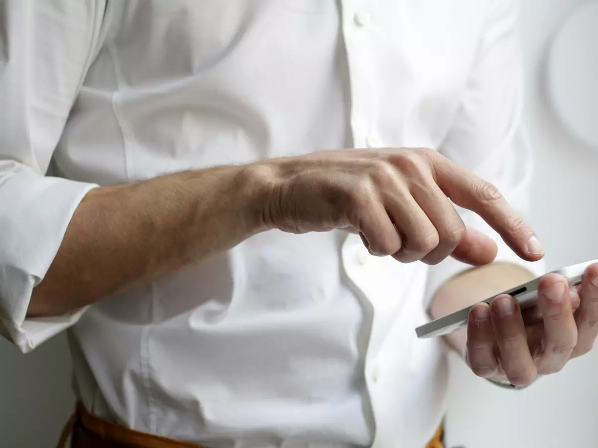 A man in a white shirt holds a white smartphone in his hands. He is interacting with the phone, using his finger to tap the screen. The phone is partially obscured by his hand, but the screen is illuminated.