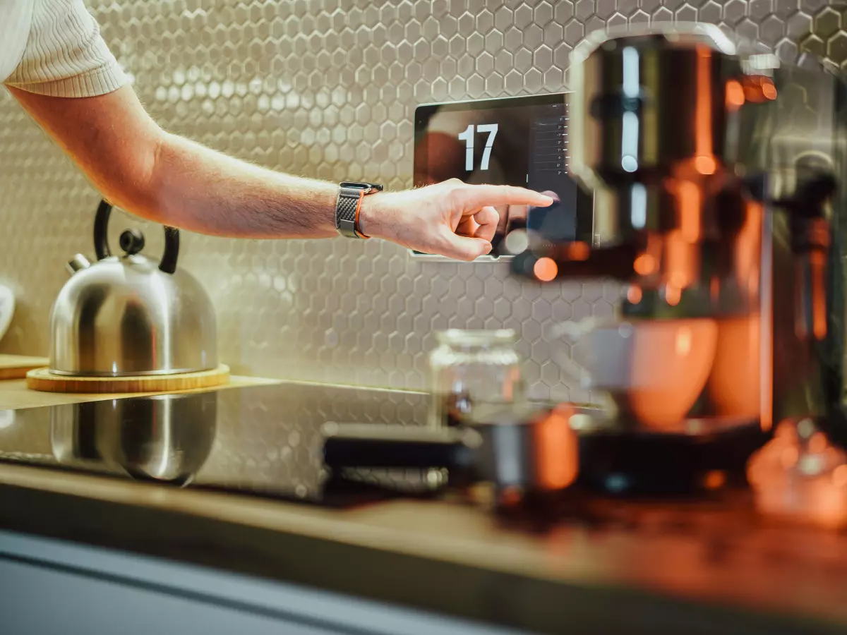 A man's hand is seen pointing at a smart screen showing a clock on a kitchen counter. There is a coffee maker next to the screen. 