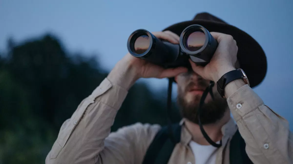 A man with a beard looking through binoculars in a natural setting at dusk.