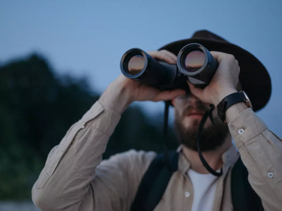 A man with a beard looking through binoculars in a natural setting at dusk.