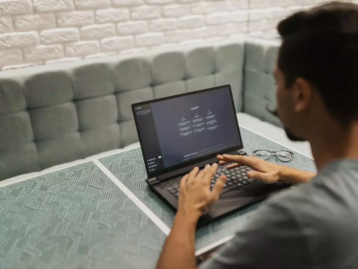 A man sitting at a table in a cafe, typing on a laptop. The screen shows code related to artificial intelligence.