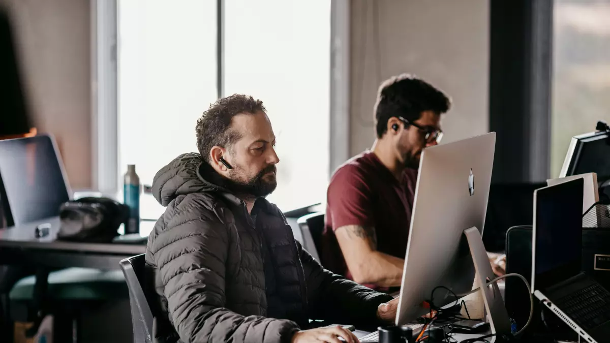 Two men working on computers in an office. One man is looking at the computer screen while the other one is typing on the keyboard. The office is modern and well-lit.