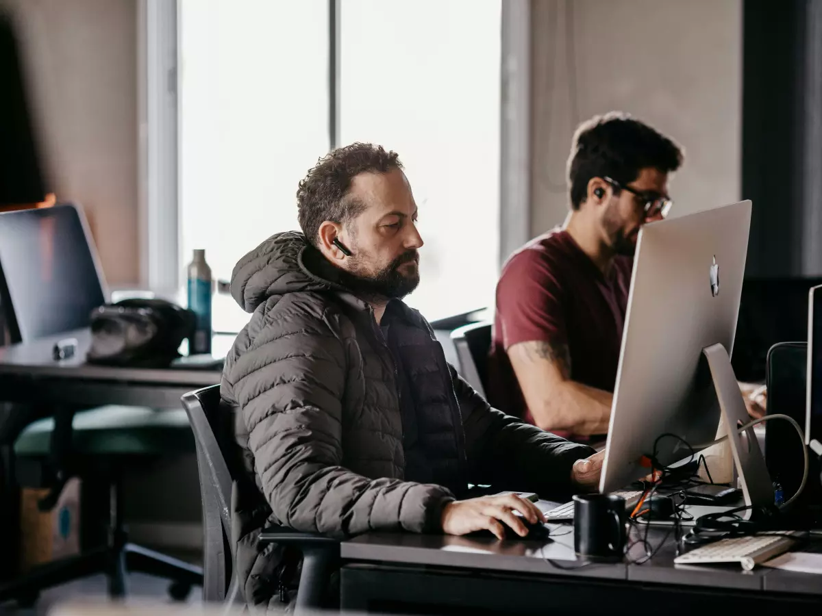 Two men working on computers in an office. One man is looking at the computer screen while the other one is typing on the keyboard. The office is modern and well-lit.