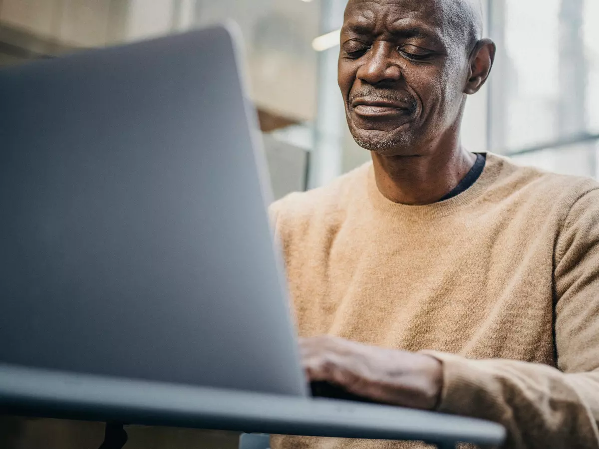 A man is sitting at a table in a modern office, working on his laptop. He is wearing a brown sweater and has a serious expression on his face.