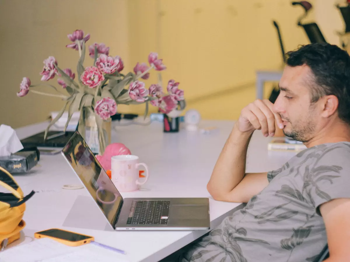 A man is sitting at a desk with a laptop open in front of him. He is leaning back in his chair with his chin on his hand. He has a serious expression on his face, but his eyes are looking towards the laptop. There is a vase of flowers and a mug on the desk, and some papers in the background.