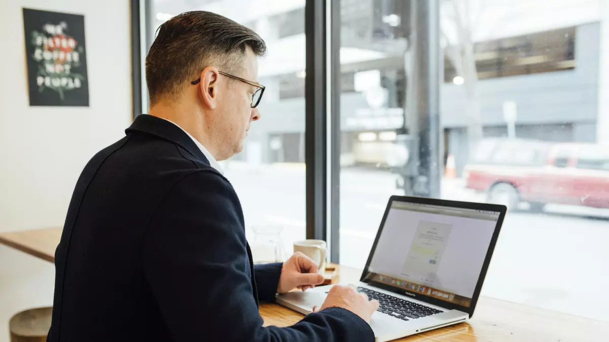 A man in a suit is working on his laptop in a cafe, looking stressed and preoccupied.