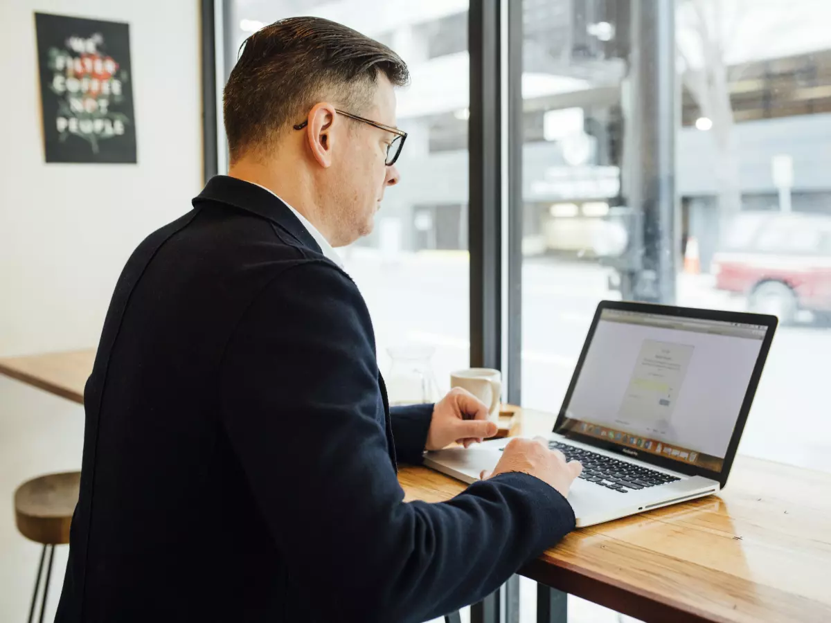 A man in a suit is working on his laptop in a cafe, looking stressed and preoccupied.