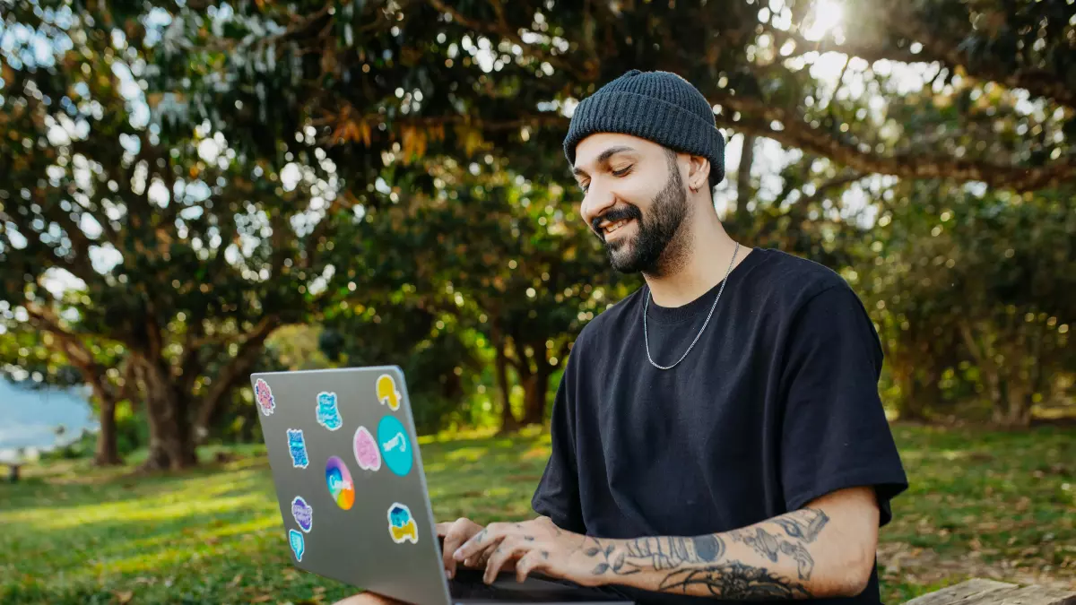 A young man with tattoos and a black beanie sits on a wooden bench in a park, using a laptop.  The laptop is decorated with stickers. 