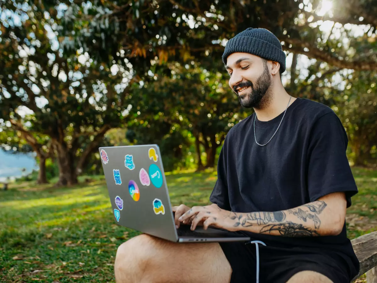 A young man with tattoos and a black beanie sits on a wooden bench in a park, using a laptop.  The laptop is decorated with stickers. 