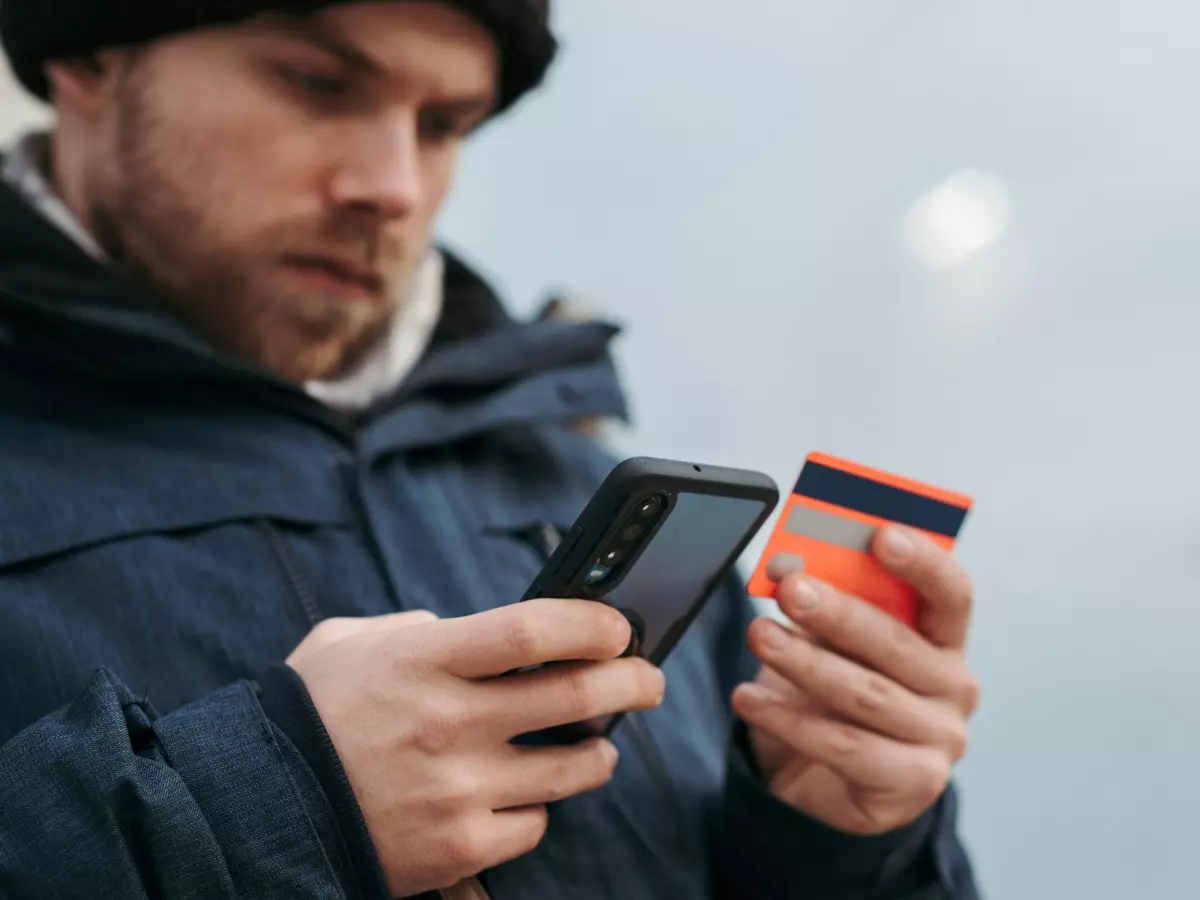 A man is holding a smartphone and a credit card in his hands. He is wearing a blue jacket and a black hat.