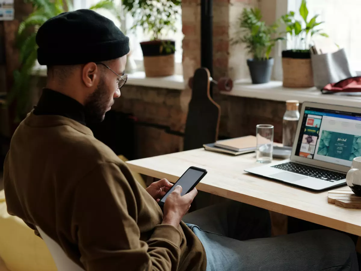 A person sitting at a desk, using a smartphone. They are wearing a brown jacket and a black beret. A laptop is open in front of them, and there are plants in the background.