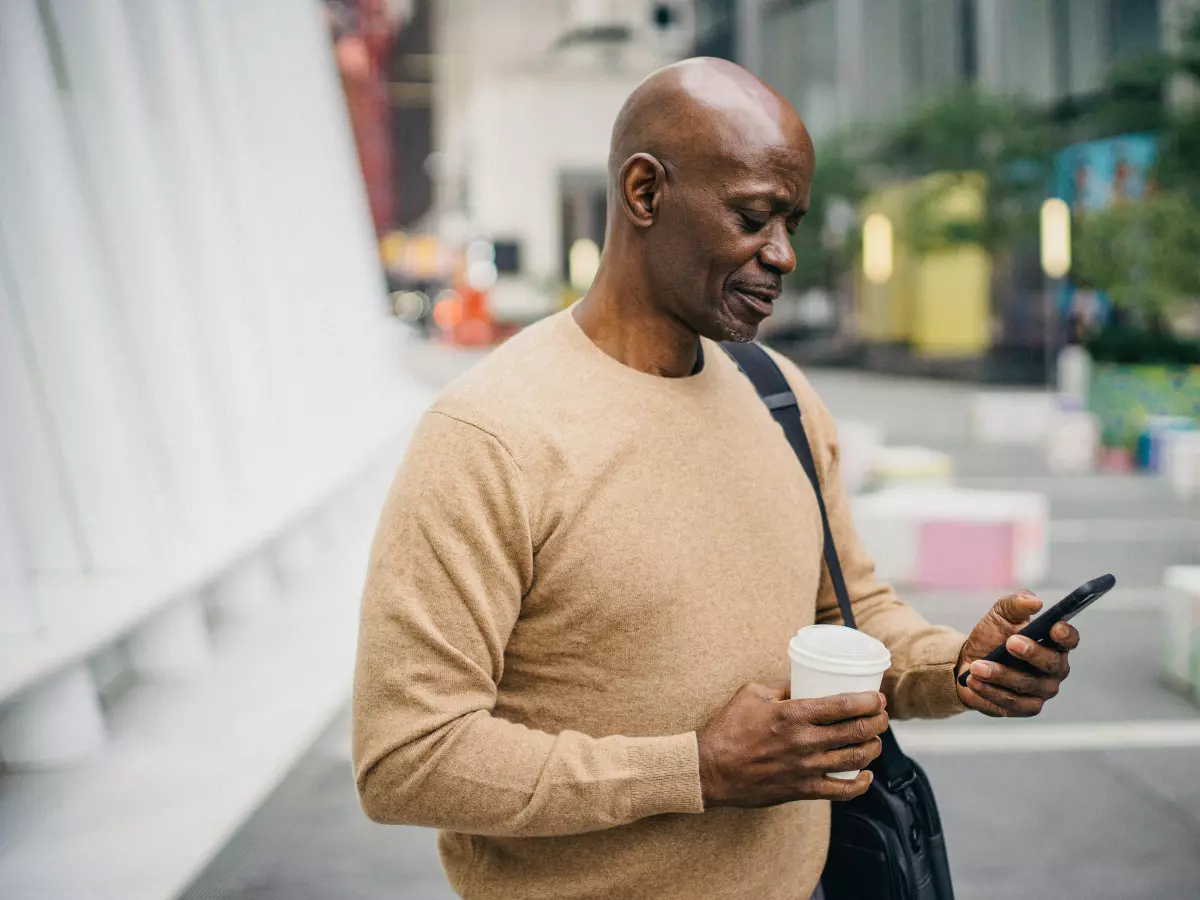 A man holding a coffee cup and a phone in his hands, looking down at the phone.