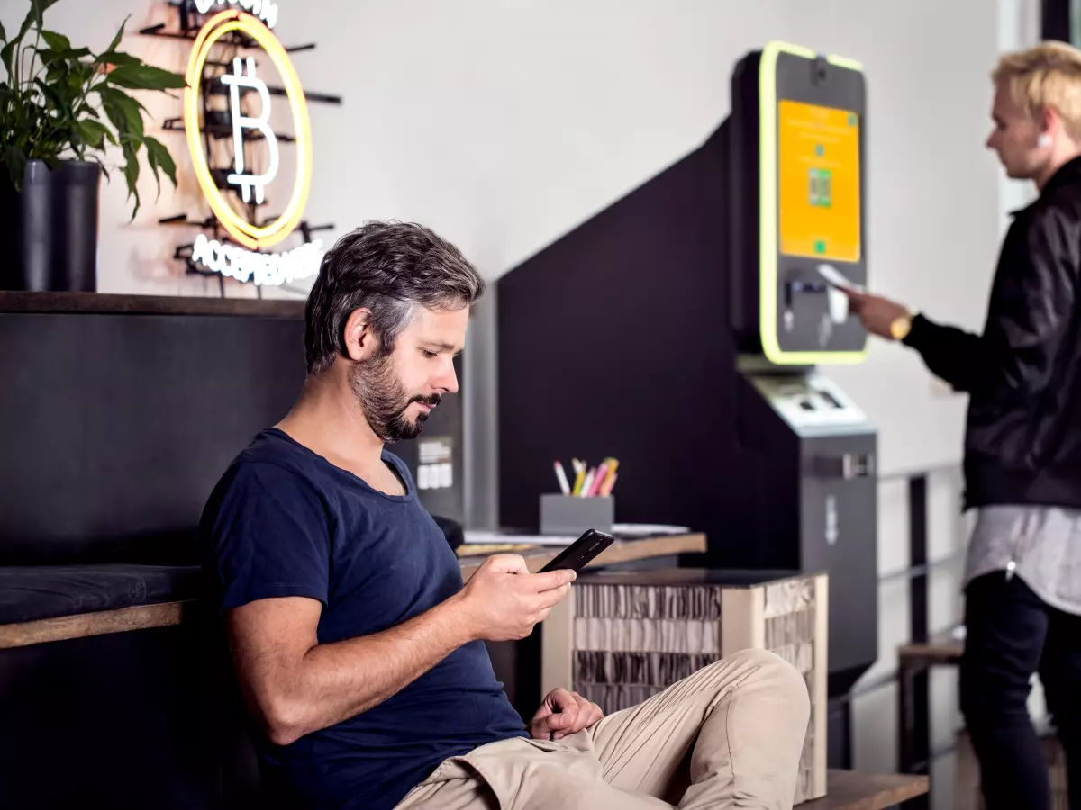 A man sitting in a cafe, looking at his phone. There is a bitcoin logo visible in the background.