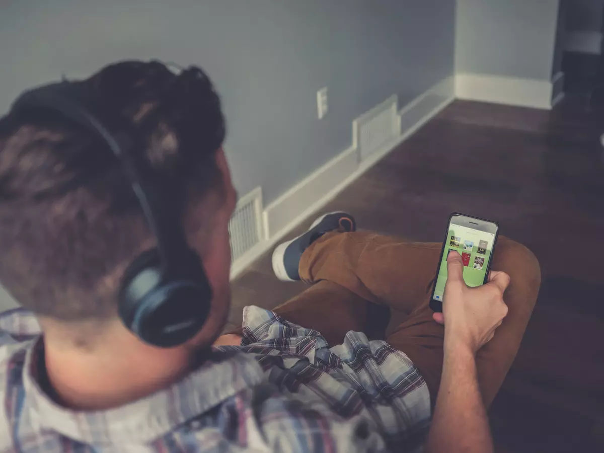 A man wearing headphones is sitting in a chair and looking at a smartphone.