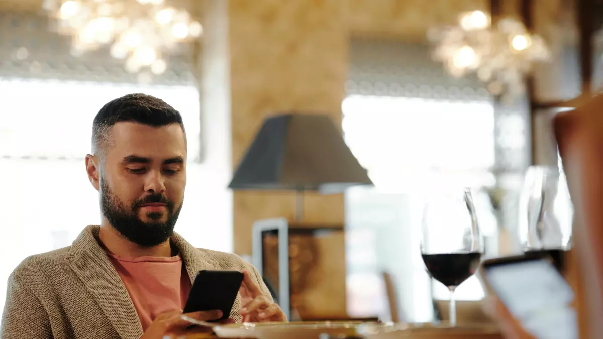 A man sitting at a table in a cafe, using his smartphone, with another person in the background.