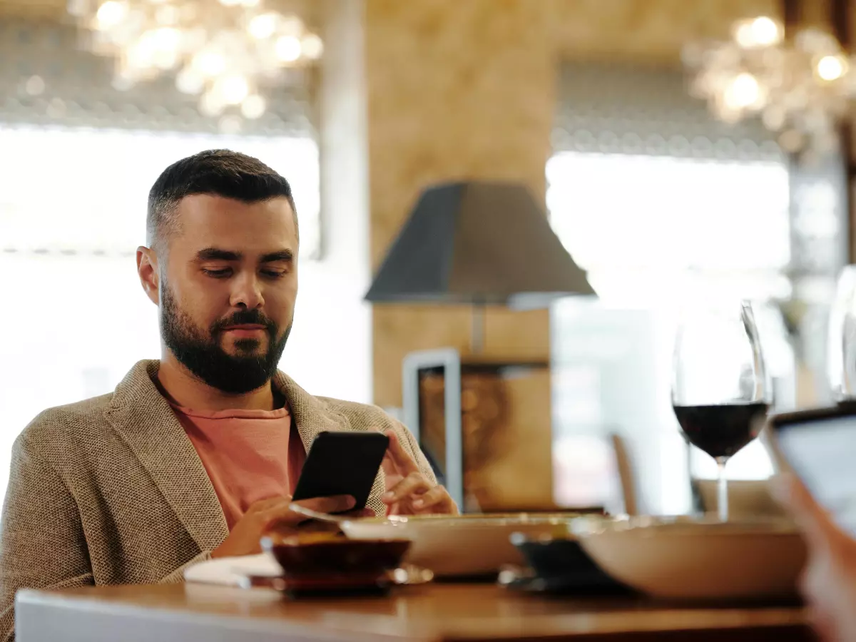 A man sitting at a table in a cafe, using his smartphone, with another person in the background.