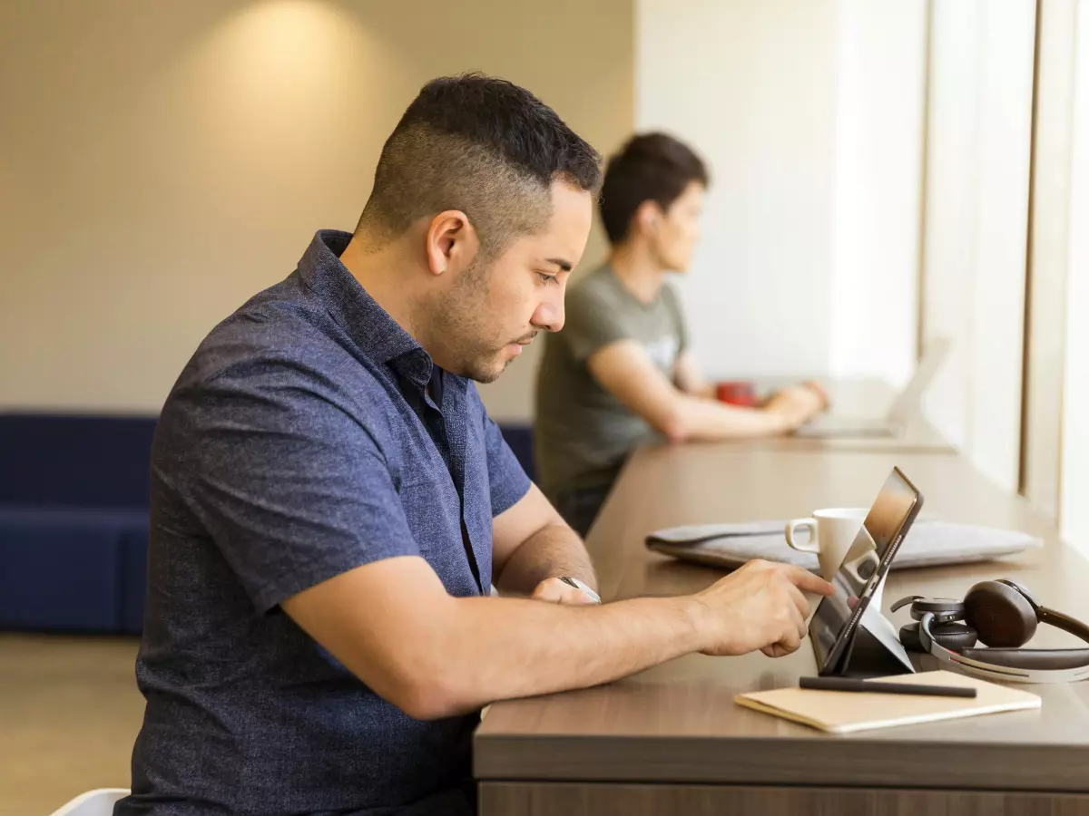 A man is sitting at a desk using a tablet. Another man is working on a laptop in the background.