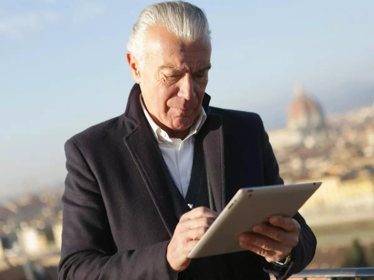 A senior man in a coat uses a stylus on a tablet with a city view in the background