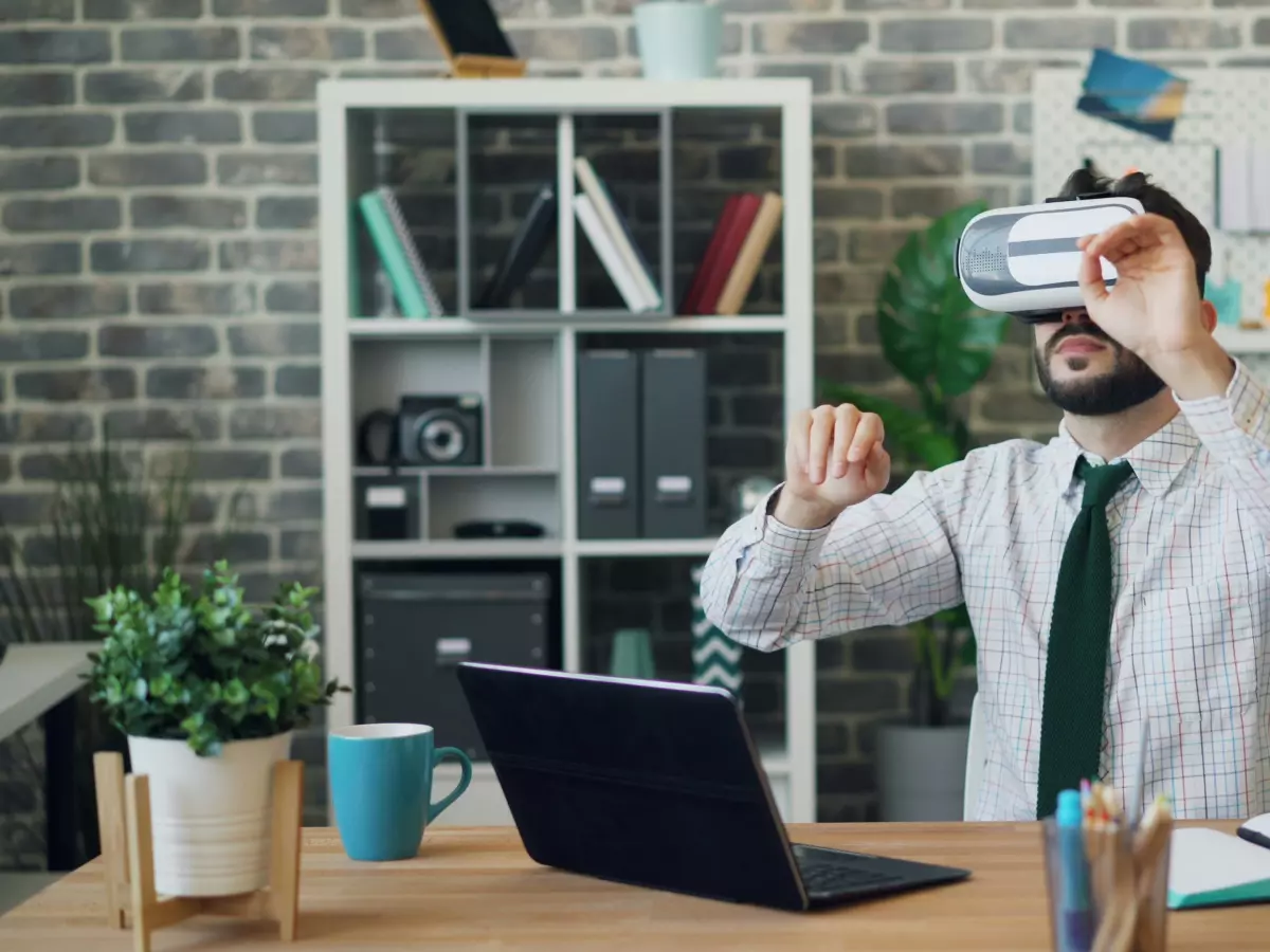 A man wearing a VR headset is sitting at a desk in an office. He is using his hands to interact with the virtual world. There is a laptop and a cup of coffee on the desk. The room is decorated with plants and a brick wall.