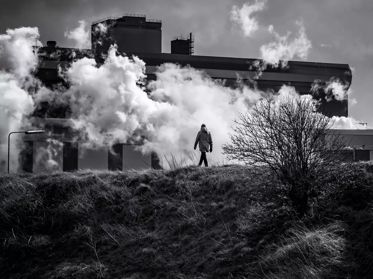 A man in a black jacket and white pants walks on a grassy hill towards a massive factory complex. The sky is cloudy and filled with dense smoke emanating from the factory's smokestacks.