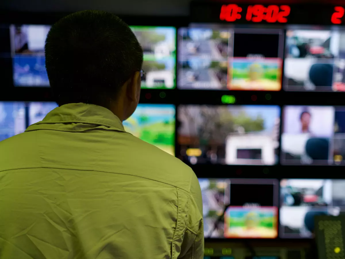 A man in a green shirt stands in a server room, watching the screens. There are multiple screens, each showing different data and information.