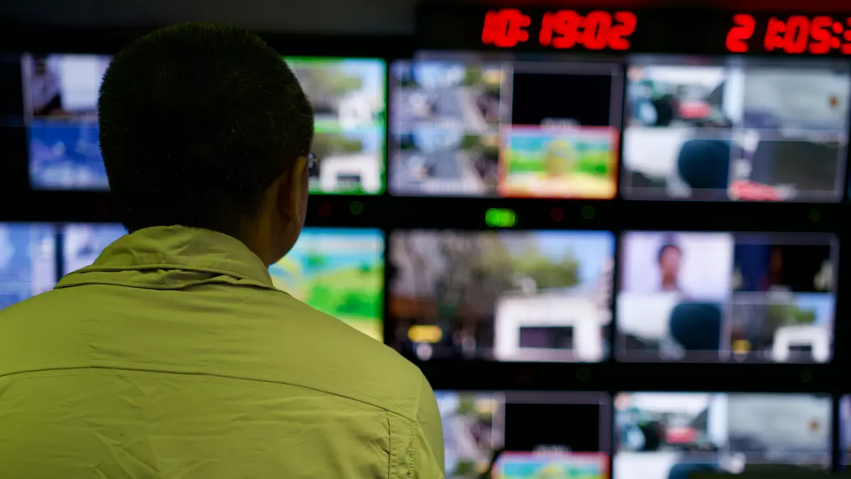 A man in a green shirt stands in a server room, watching the screens. There are multiple screens, each showing different data and information.