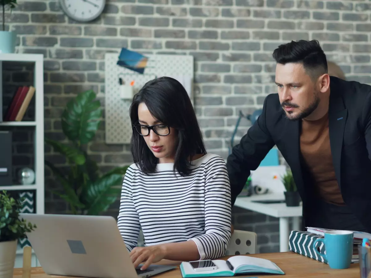 Two people working together on a laptop in an office setting. The woman is typing on the keyboard, and the man is looking at the screen.