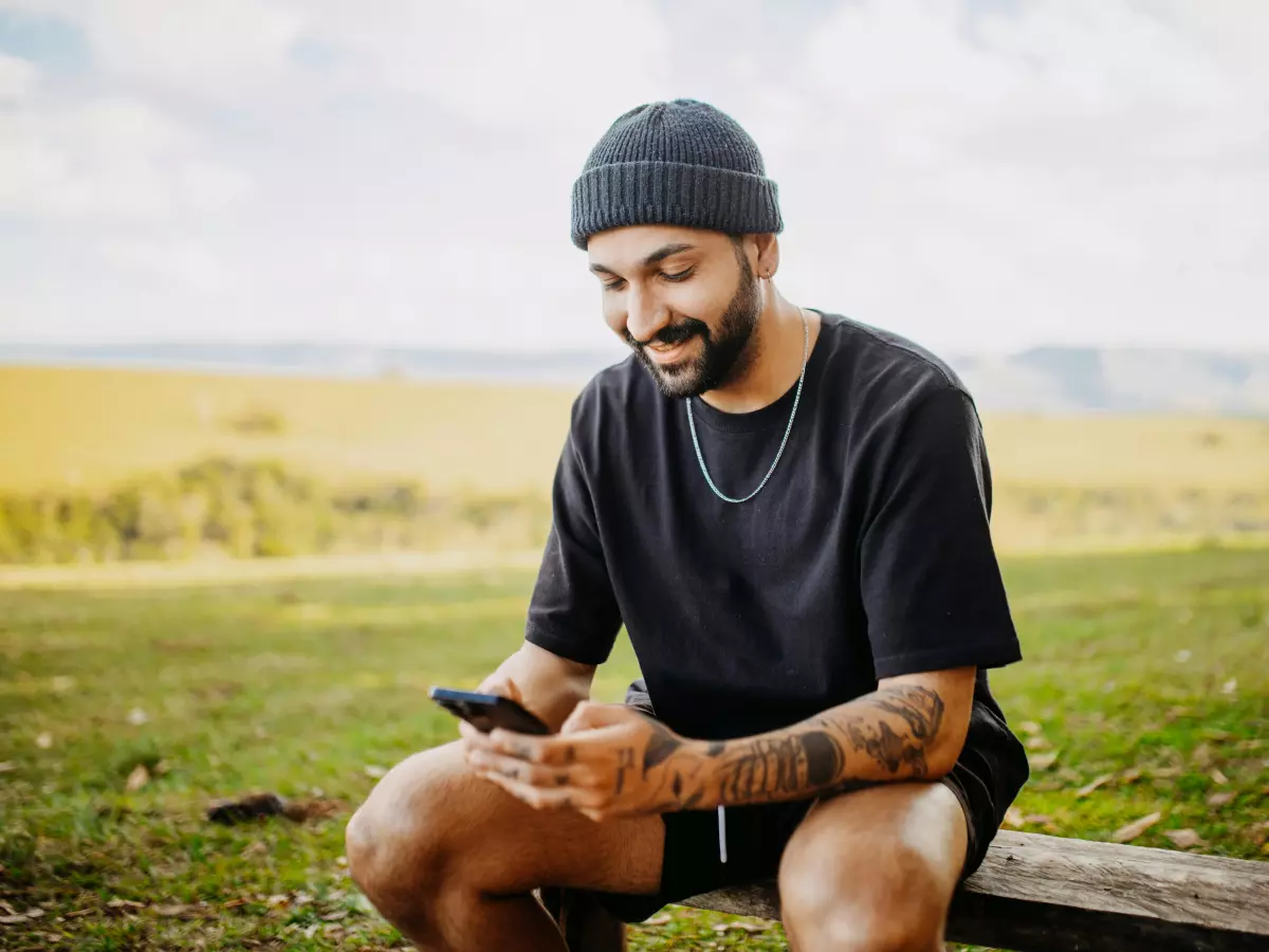 A man sitting on a wooden bench, looking at his smartphone with a frustrated expression. The man is wearing a black t-shirt, black shorts, and a black beanie. He has a beard and tattoos on his arms. The background is a field of green grass and a blue sky with white clouds.