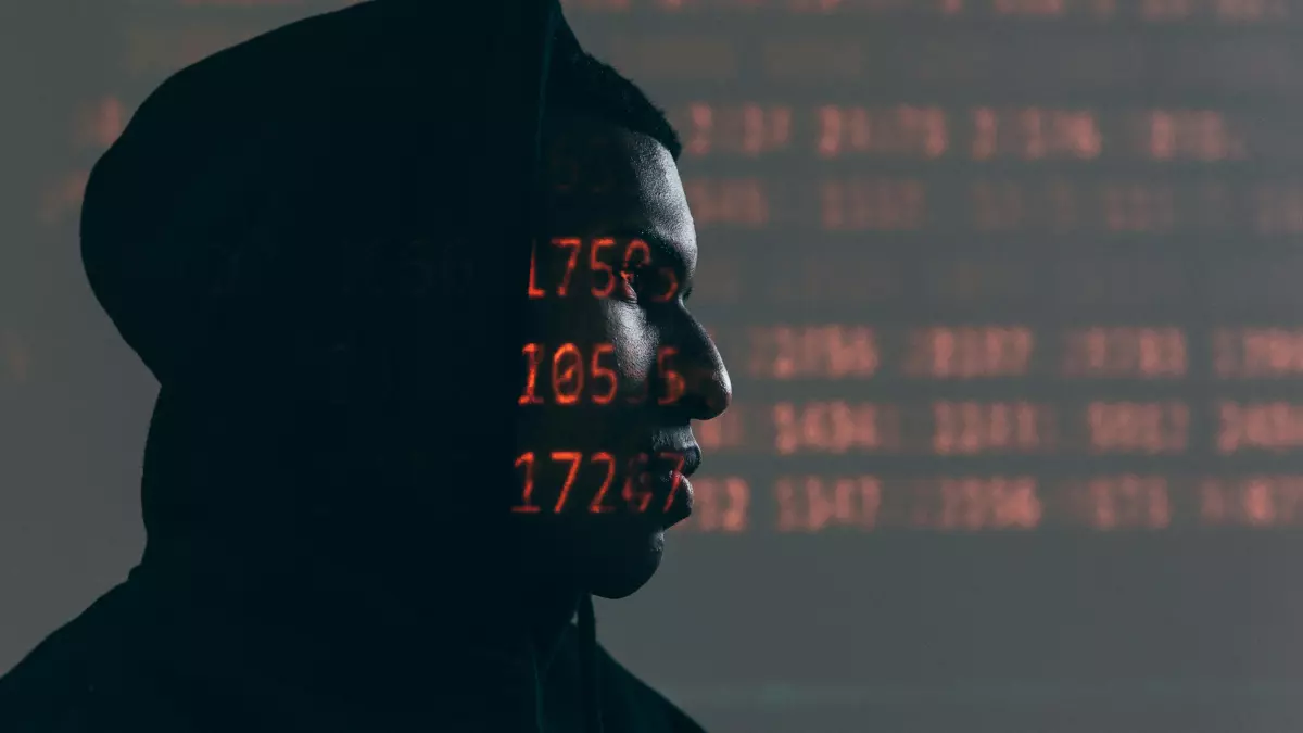 A person wearing a hoodie with their face partially obscured, standing in front of a screen displaying code.