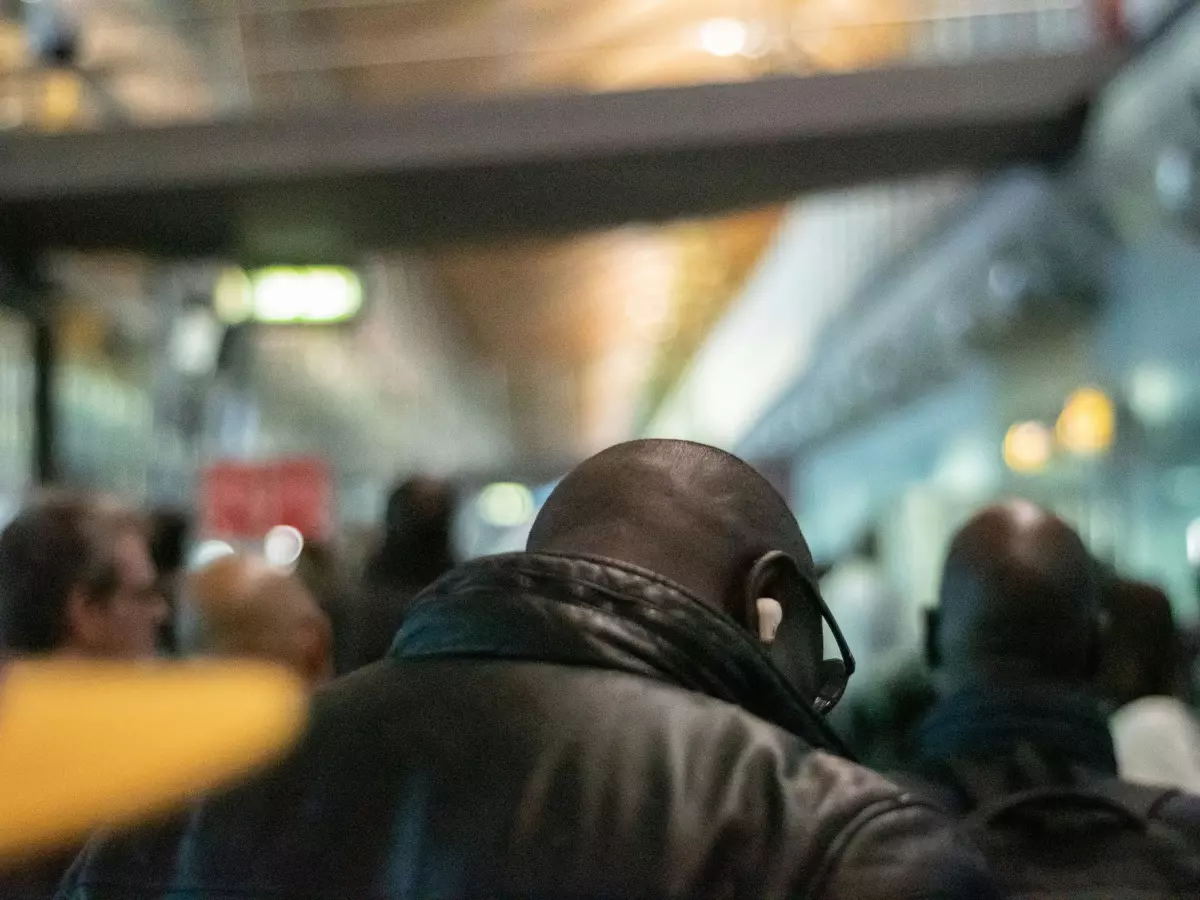 A man with headphones on walks through a crowded airport. He is in the background, and the focus is on the headphones.