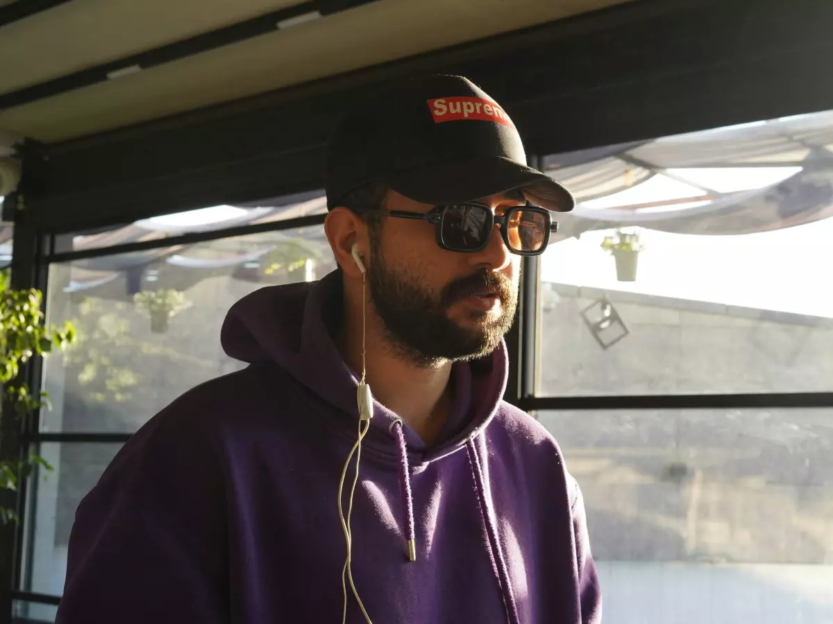 A man wearing a black hoodie and sunglasses listens to music with white earbuds in a urban setting.