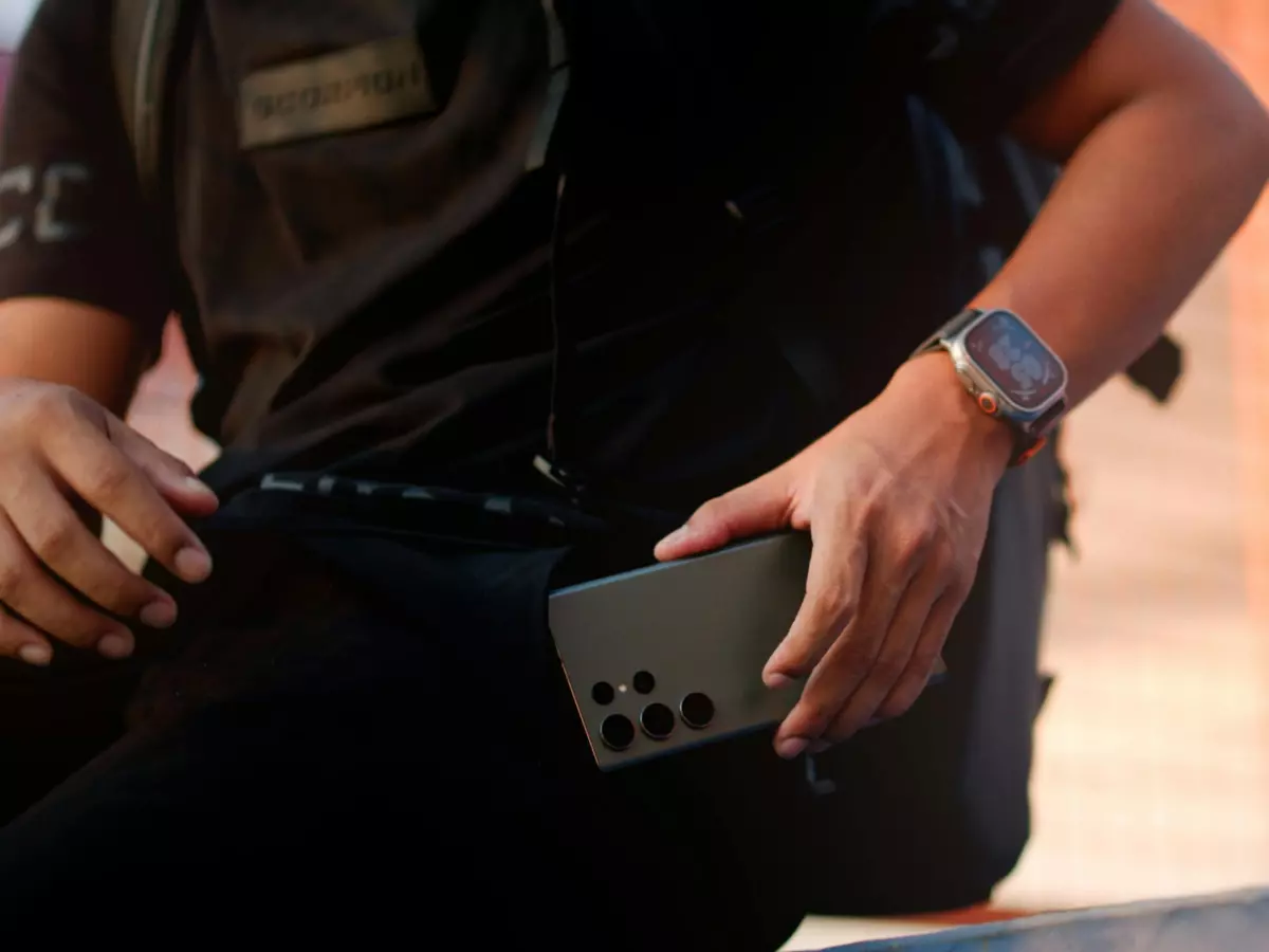 A person wearing a black t-shirt and black pants sitting on a staircase with a black Galaxy S24 FE in their hand and a silver Galaxy Watch FE LTE on their wrist.