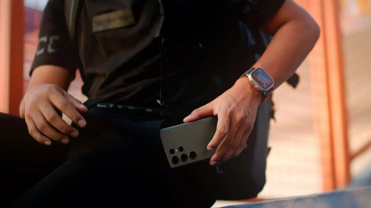 A person wearing a black t-shirt and black pants sitting on a staircase with a black Galaxy S24 FE in their hand and a silver Galaxy Watch FE LTE on their wrist.